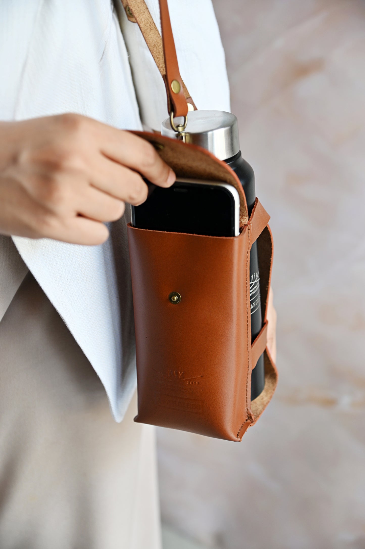 Brown handcrafted leather bottle case with utility pocket and steel flask, worn crossbody