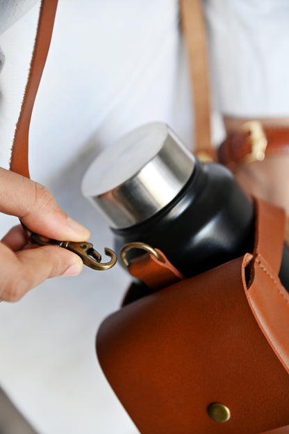 Brown handcrafted leather bottle case with utility pocket and steel flask, worn crossbody